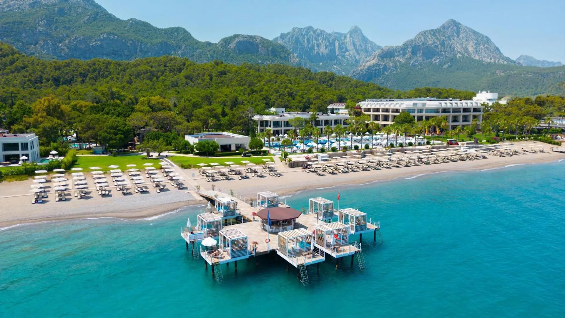 Sherwood Exclusive Kemer - Kids Concept Goynuk  Exterior photo The photo shows a beautiful coastal resort scene on a sunny day. The foreground is dominated by a stunning turquoise sea, calm and inviting.  A long wooden pier extends out into the water, featuring several open-air cabanas or lounge areas with seati
